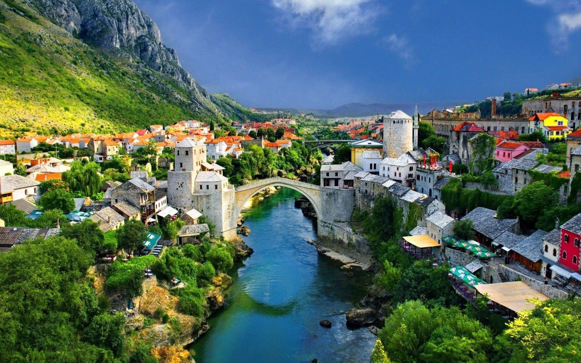 Townscape with a river and bridge, surrounded by greenery and mountains under a blue sky.