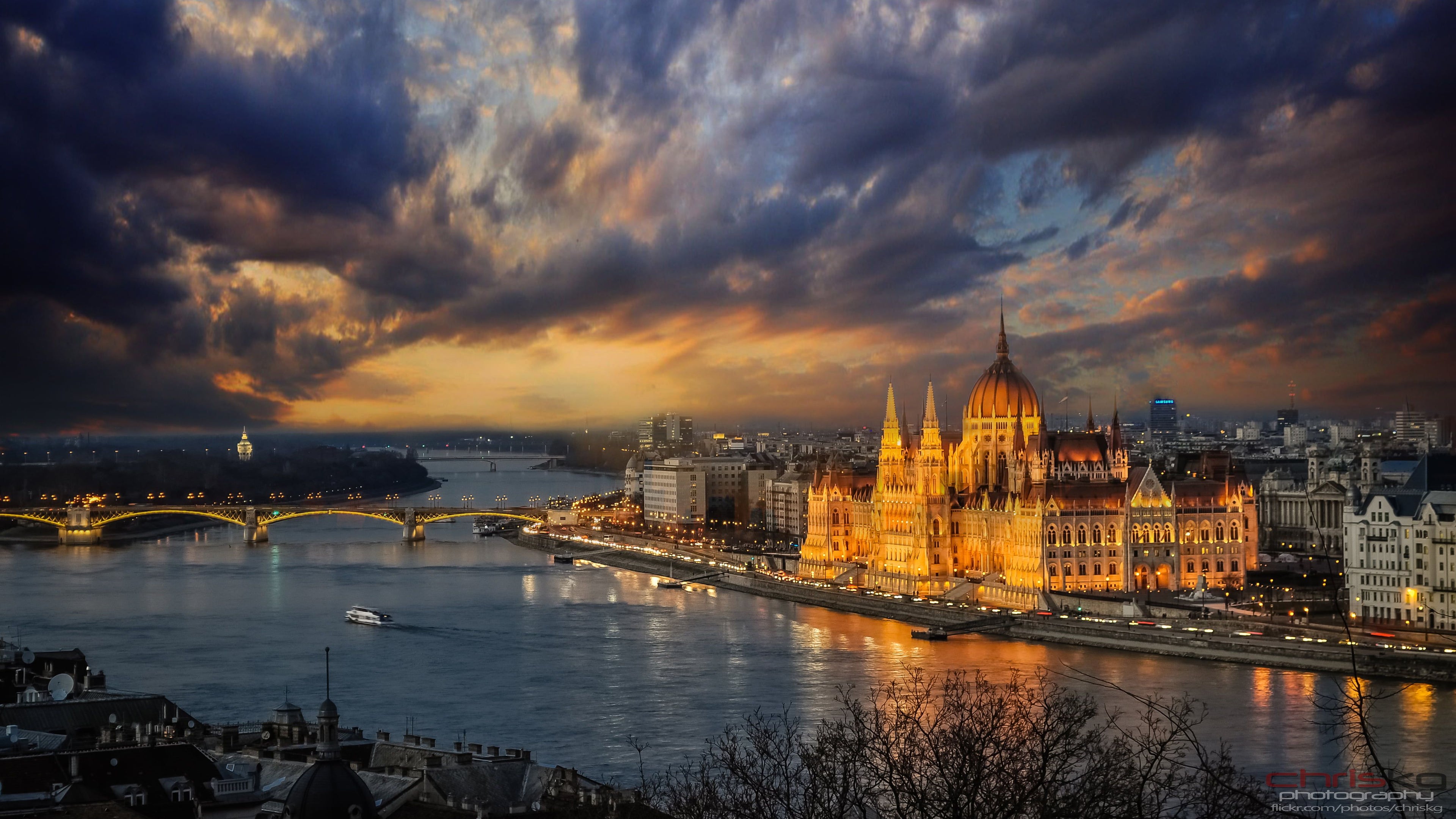 Cityscape with illuminated buildings and a river under a dramatic sky.