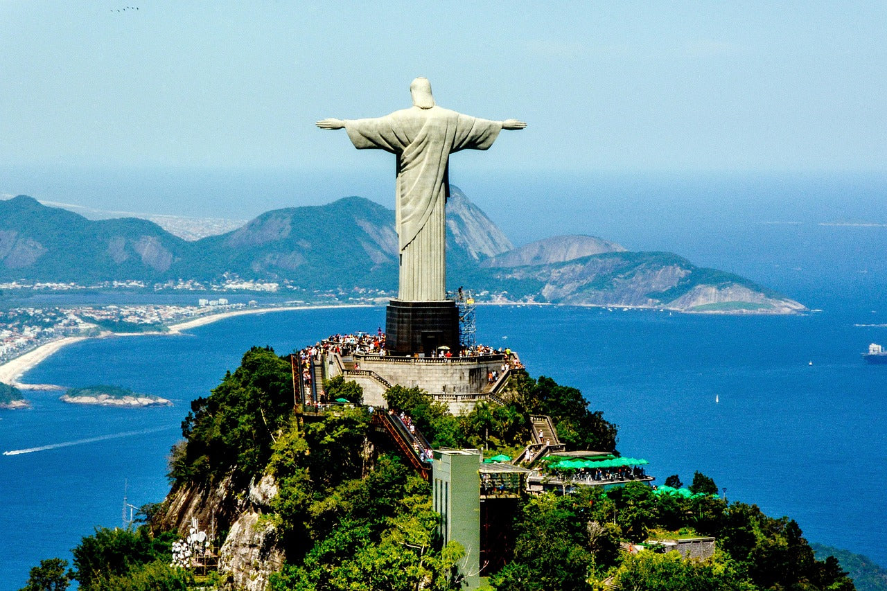 Statue of Christ the Redeemer overlooking a coastal city with mountains and blue sky.