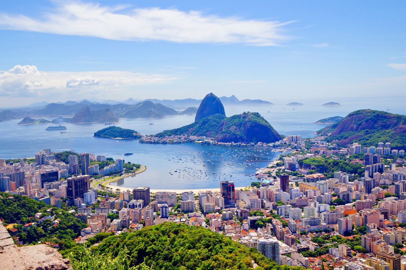 Cityscape with buildings, a body of water, and a mountain under a blue sky.